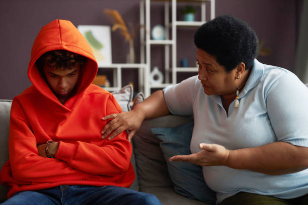 African American mature woman talking to sulky teenage grandson African American mature woman talking to sulky or stressed teenage grandson in red hoodie while sitting on couch next to him gloomy mood stock pictures, royalty-free photos & images