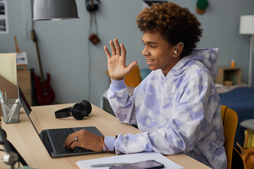 Happy schoolboy in hoodie greeting teacher on laptop screen by waving hand in the beginning of online lesson while sitting in bedroom