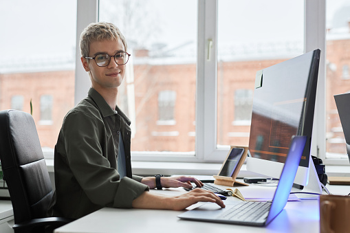 Portrait of young programmer in eyeglasses looking at camera while developing software on computer at his workplace