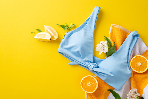 Stylish summer vacation concept. Top view flat lay of blue swimwear, orange lemon sunglasses, striped towel and flowers on bright yellow background with empty space for text or advert