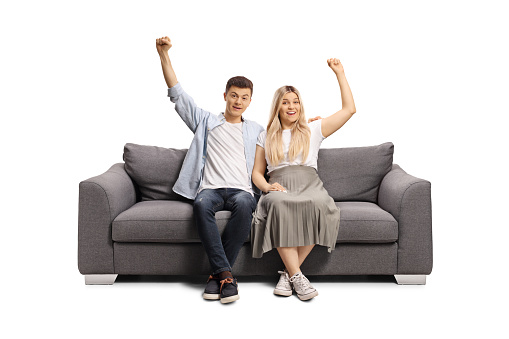 Cheerful young couple sitting on a sofa and raising arms isolated on white background