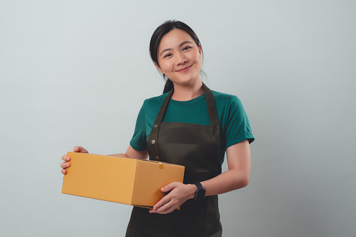 Asian woman wearing apron happy smiling holding carton box and looking at camera, standing isolated on background.