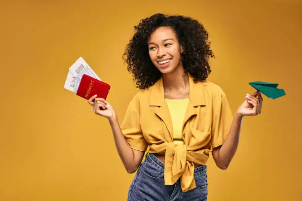 Air travel concept. Smiling curly African woman with air tickets and international passport throws paper airplane simulating air traveling, over yellow background