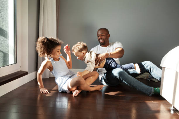 Happy Young Black Family Playing Together On Floor At Home In Living Room. Funny dad having fun with two kids. African american father with children, real people emotion stock photo