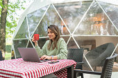 Woman  Sitting At Table And Using Laptop In Front Of A Geo Dome Glamping Tent.
