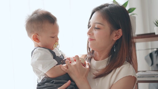 Kind asian mom sitting at cozy home carry and embracing her cute baby boy. Parenting and attentiveness for growth and good development of children, moments of happy mother. Concept of family, motherhood, life insurance.