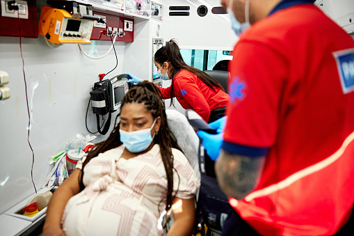 Uniformed man and woman monitoring expectant mother’s blood pressure before transporting her to hospital, everyone in protective face masks.
