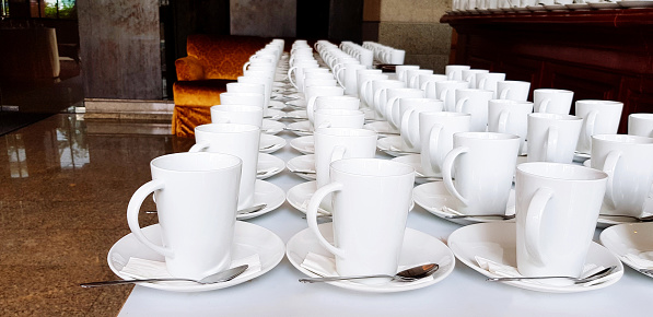 Many cup of coffee or tea and stainless steel spoon putting on white table for serving customer and staff in hotel or Ballroom. Group of object, Food, Drinking, Service and Preparation concept