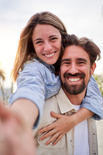 Happy blonde couple using smartphone to take a selfie while doing piggyback, looking at camera smiling enjoying summer trip outside. Vertical photo.