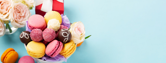 Sweet cookies macaroons, red gift box, rose flowers and chocolate candies on blue background. Spring presents concept for Mothers day, womans day, birthday. Banner, empty space for text.