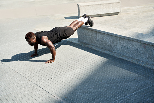 Fit sporty young black man doing push ups exercises in urban park. Strong muscular healthy African ethnic guy bodybuilder doing sport fitness street workout training in outdoor city gym.