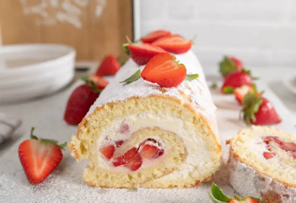 Delicious strawberry cake or dessert with traditonal swiss roll filled with whipped cream and marinated strawberries. Served ready to eat with cross section view on light kitchen counter. Front view