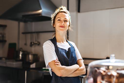 Female cafe owner, showing confidence in her small business, stands in front of the camera with arms crossed. Woman wearing an apron, proudly displays her role as a hospitality entrepreneur.