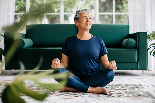 Happy senior woman practicing yoga in the lotus position, meditating for inner peace and mindfulness. Mature woman doing a breathing exercise in the tranquility of her home, sitting on the floor in fitness clothing.