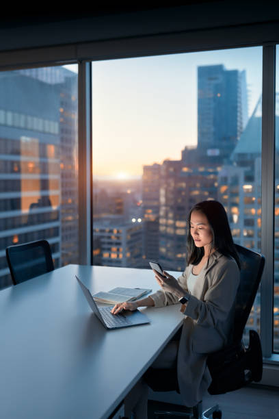 Young busy Asian business woman working using mobile at night in office. Young busy Asian business woman using laptop and mobile cell phone tech at night in dark office. Professional businesswoman holding smartphone, working on cellphone with evening city view. Vertical office window night stock pictures, royalty-free photos & images