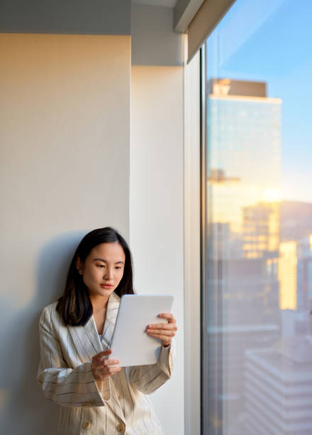 istock Young Asian business woman using digital tablet standing at office window. 1487464893