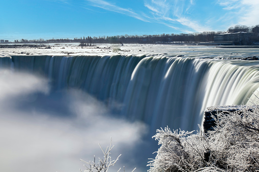 Panoramic view over the Niagara Falls, ON, Canada during winter with smooth slow shutter speed water flow from the falls and white ice on the bushes and vegetation as result of water spray