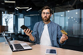 Portrait of disappointed and deceived businessman inside office, mature man looking at camera confused holding bank credit card and phone, employee got rejection error