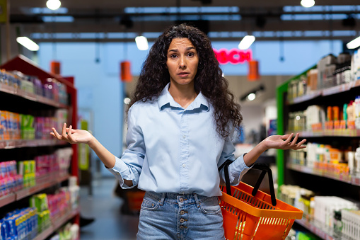 A worried young Latin American woman stands in a supermarket between the shelves with goods, holds a basket, spreads her hands, looks at the camera. Unable to find product, out of stock.