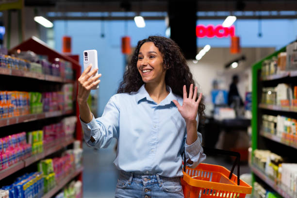 achats. une jeune femme latino-américaine marche dans un supermarché entre des rangées d’étagères avec un panier. conversations téléphoniques, appel vidéo - supermarket discussion people talking photos et images de collection