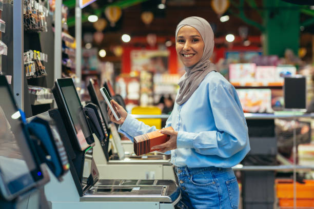 selbstbedienung im supermarkt. eine junge arabische frau im hijab steht in der nähe der elektronischen registrierkassen und bezahlt die waren im geschäft online über eine anwendung auf ihrem handy - turkish ethnicity white black contemporary stock-fotos und bilder