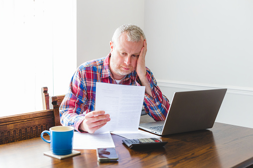 Portrait of a mid adult man in his 30s checking his energy bills at home. He has a worried expression and touches his face with his hand while looking at the bills. He is dressed casually in a check patterned shirt.