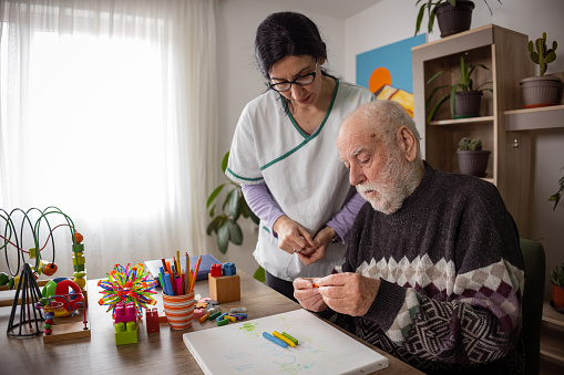 A kind nurse helps a senior man with dementia express his creativity through drawing with colorful crayons.