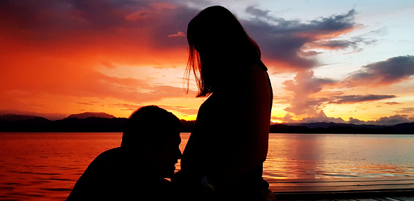 Silhouette of couple lovely at sunset time. Husband kiss on wife's belly there has baby inside with orange sky, cloud and lake background. Lover of family or parents and Pregnant concept
