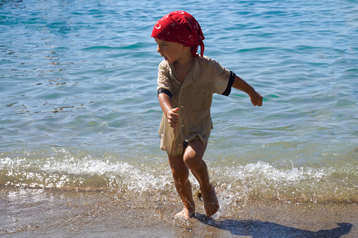 A little boy in a red bandana runs out of the sea. Blurry motion and splashes. The little ones are merrily running along the ocean. Summer family vacation.