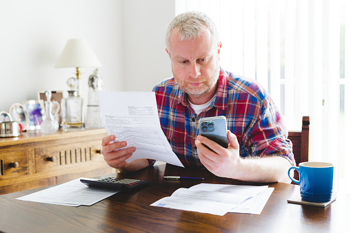 Portrait of a mid adult man in his 30s checking his energy bills at home. He is using his smart phone device to pay the bills. He is dressed casually in a check pattern shirt while sitting at a table in his living room.