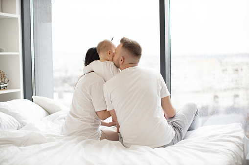 Rear view of loving father kissing his baby daughter while little girl hugging caring mom in morning after waking up. Young married couple with kid resting on white blanket in bedroom of modern flat.