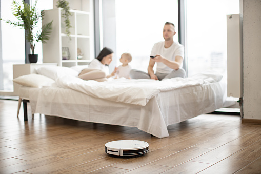Brunette woman playing with infant girl on bed while young man turning on robot vacuum cleaner via remote control in studio room. Modern family resting at weekend using one touch home cleaning.