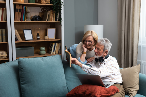 Simple living. Happy middle-aged senior couple 70s husband and wife sit relax on couch in living room reading book drinking tea together, calm elderly 60s couple booklovers rest at home enjoy life