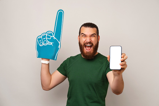 Ecstatic man wearing green t shirt and foam finger fan glove while showing blank phone screen.