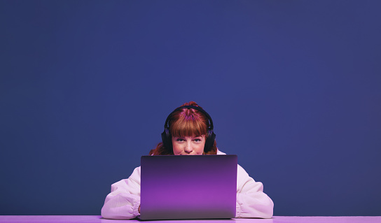 Woman with ginger hair looking at the camera while sitting behind her gaming laptop in a studio. Female gamer using an interactive streaming service while sitting in neon purple light.