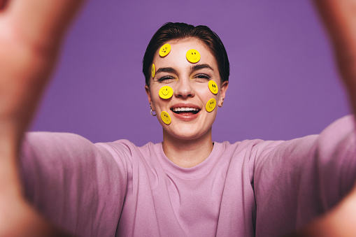 Playful young woman taking a selfie with smiley stickers on her face. Happy woman smiling at the camera while taking a picture of herself. Cheerful young woman having fun against a purple background.