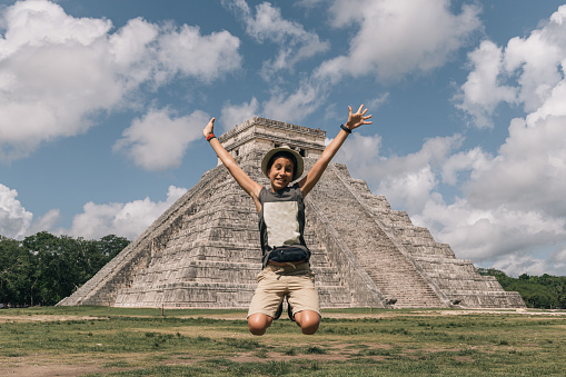 Happy teenage girl with hat jumping with open arms next to the pyramid of Kukulcan in Chichen Itza, Yucatan, Mexico