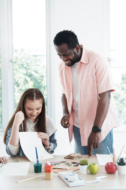 istock Young African teacher or tutor is holding book and teaching for knowledge about Art and watercolor painting and drawing for Caucasian student girl in classroom. Education and artist Concept. 1487494373