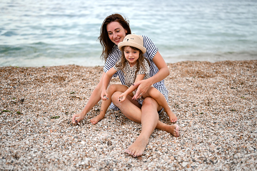 woman sitting with her daughter on a seashore and playing together with the kid.
