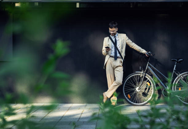 Portrait of business man with bicycle standing in front of office building looking at smartphone. stock photo