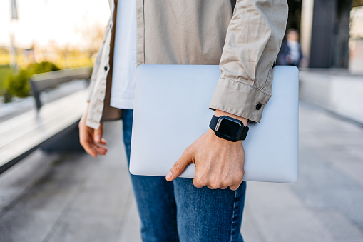 Young man wearing a smart watch and holding a laptop while walking in front of a corporate building outdoors. Close-up.