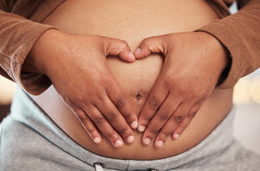 Pregnant woman, closeup of heart hands on belly and happy black mother alone in Jamaica living room. Excited future parent, holding healthy abdomen at home and natural beauty in pregnancy care