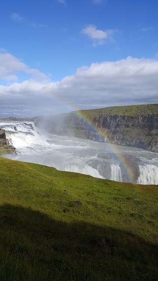 Colorful rainbow over iclandic landscape with a typical waterful foss in green grass under cloudy blue sky