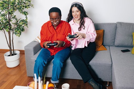 A young African American woman and a young Caucasian woman are cheerfully playing console in their living room.