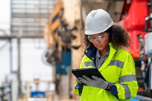 Working with a smart and confident. A female African engineer is working at a factory and examining machinery facility by a digital tablet.