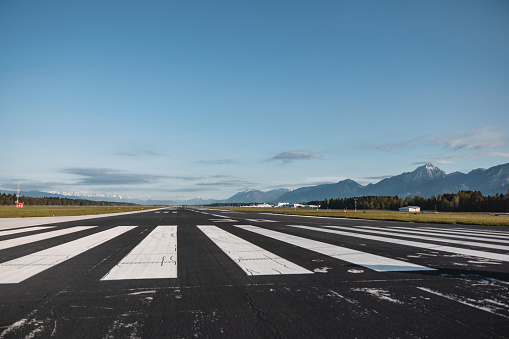 Portrait of an airstrip in the middle of airport. There are white lines on road. There are mountains in the background.