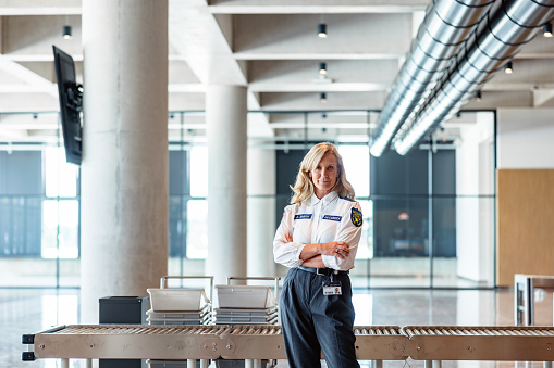 Medium shot portrait of an adult Caucasian female security officer smiling with closed mouth and looking at the camera. She is standing in front of a roller conveyors table with crossed arms. There is no travellers in the back, the airport is empty.