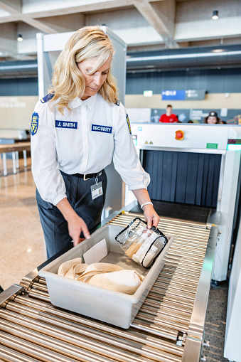 Adult Caucasian blonde female airport security officer checking the tray before pushing it inside the x-ray scanning machine.