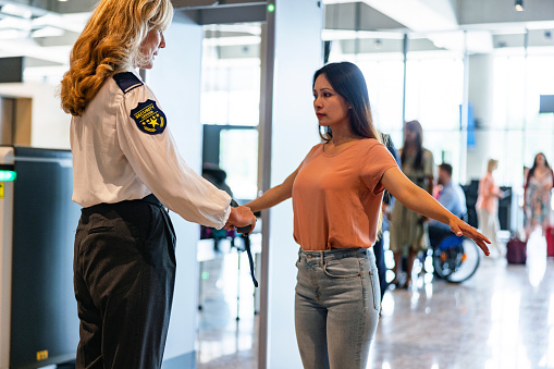 A friendly blonde airport staff member in uniform conducts a security screening on a casually dressed young woman with long hair, ensuring a safe and pleasant travel experience.