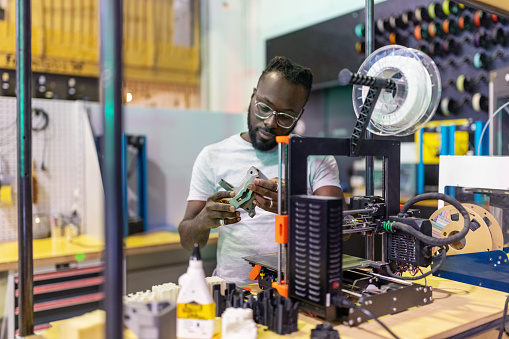 Young African man examining a 3d printed part while standing by 3D printing machine at creative lab. Male engineer using 3D printer to print a part in makers space workshop.